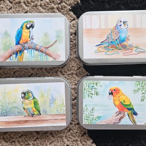 4 metaldåser med forskellige motiver af henholdvis blågul ara der sidder på en pind og tygger på garn, en blå undulat der er filtret ind i regnbue garn, en grønkindet conure med en maskemarkør i munden og en solparakit med en maskemarkør, der ligner et hjerte, i munden.